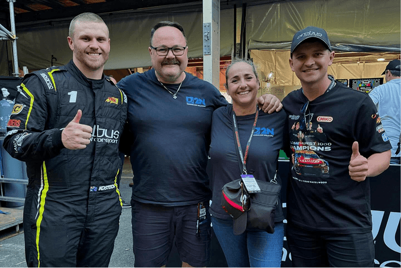 Four people posing together at an event, with two of them wearing racing suits.